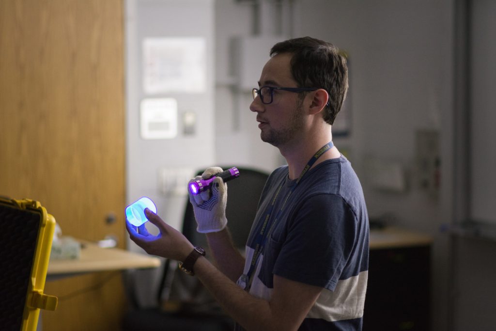 A person in a blue and white striped t shirt holding a purple flashlight to a cylindrical object in their other hand